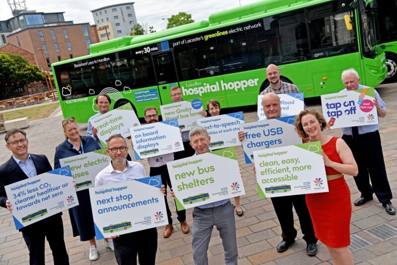 Group of people holding signs promoting Leicester's Hospital Hopper bus features like free USB chargers and next stop announcements.