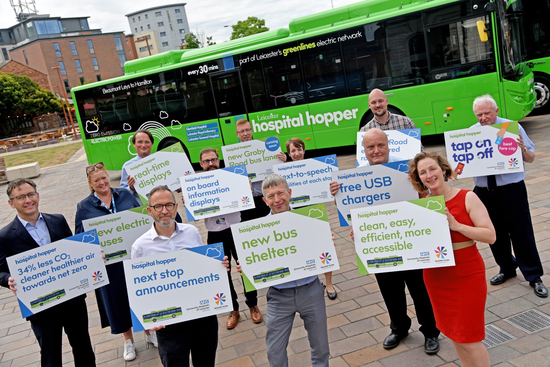 Group of people holding signs promoting Leicester's Hospital Hopper bus features like free USB chargers and next stop announcements.