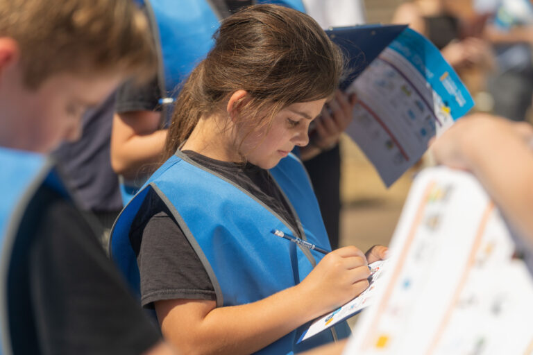 A young girl wearing a bright blue vest and black t-shirt concentrates on filling out a form with a pencil.