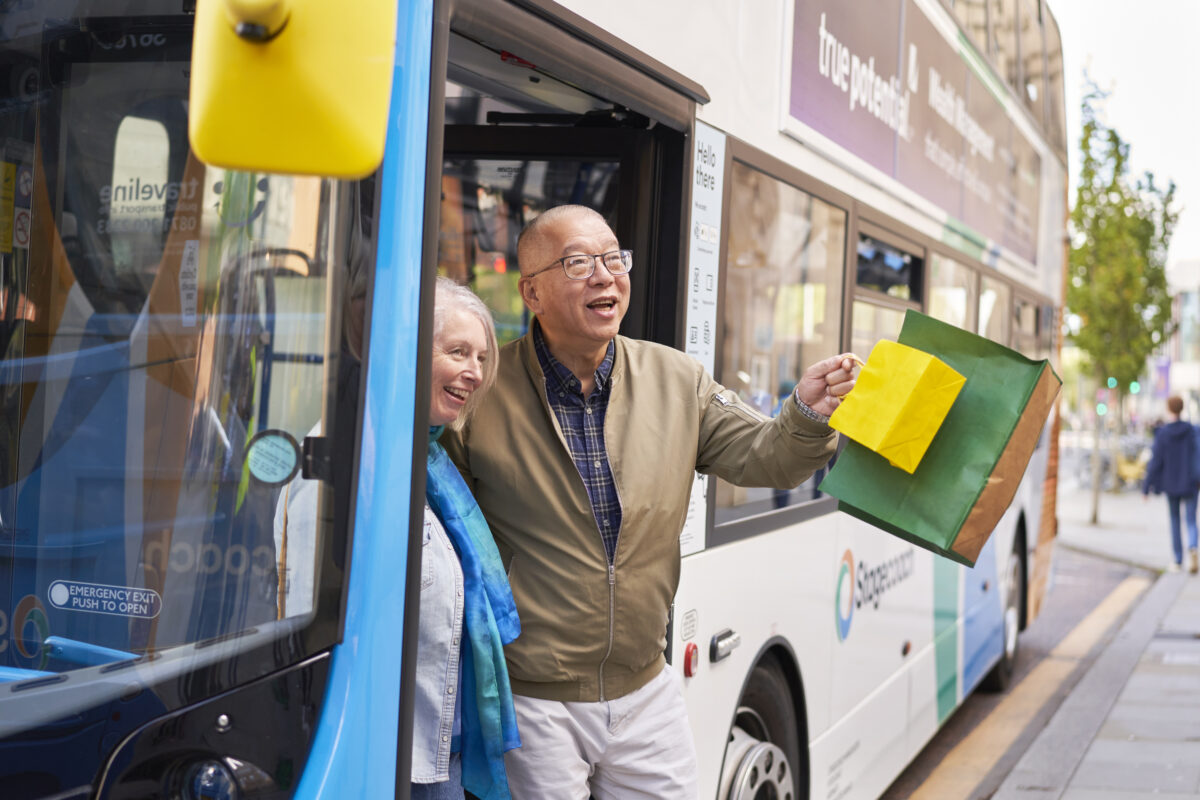 Man stepping off a city bus holding colorful shopping bags in one hand while wearing a beige jacket and plaid shirt.