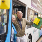 Man stepping off a city bus holding colorful shopping bags in one hand while wearing a beige jacket and plaid shirt.
