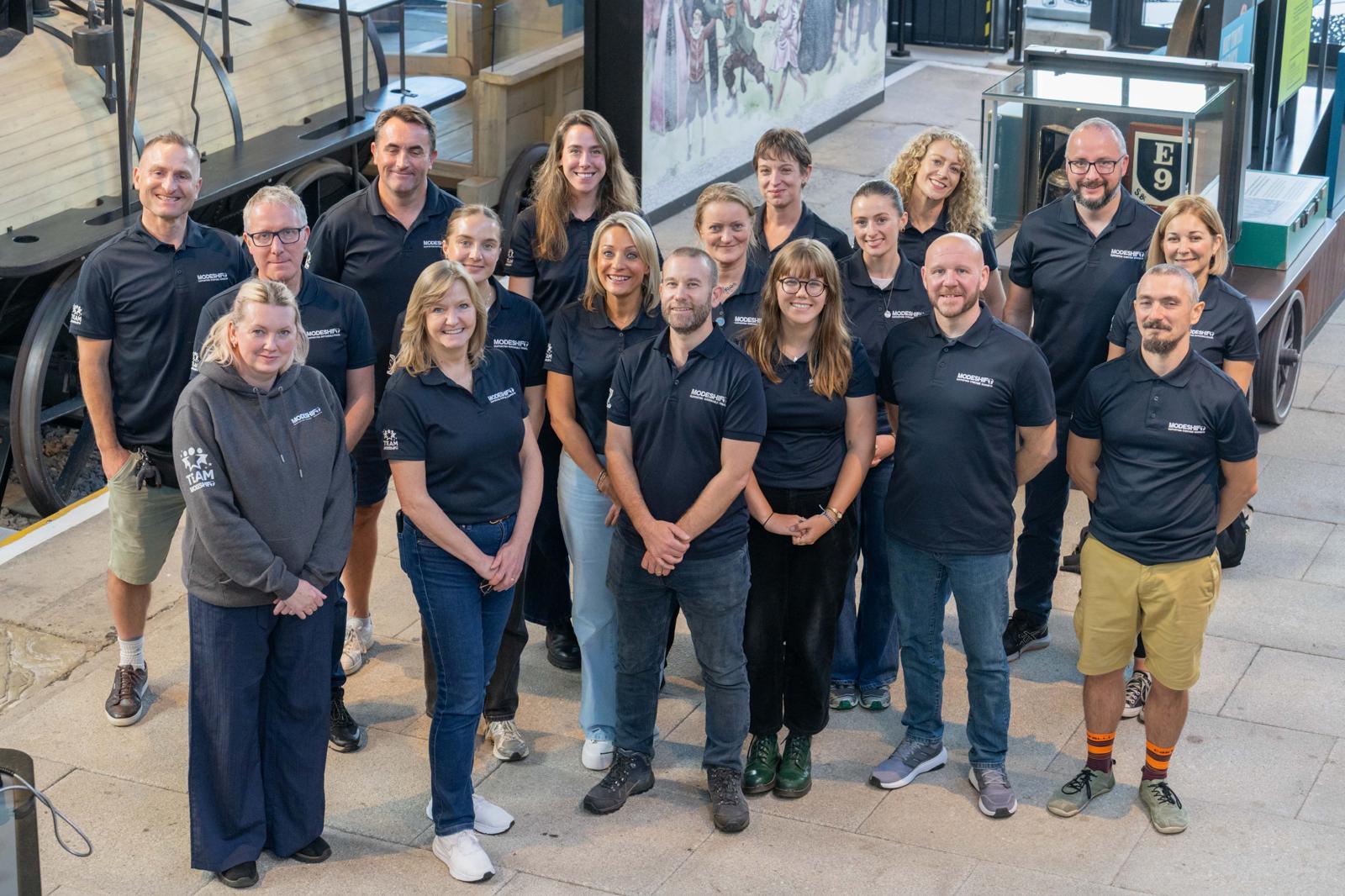 A group of people wearing Modeshift branded tops smile for the camera.