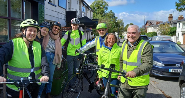 Group of cyclists wearing bright yellow safety vests standing with their bikes on a sunny street lined with parked cars and buildings.