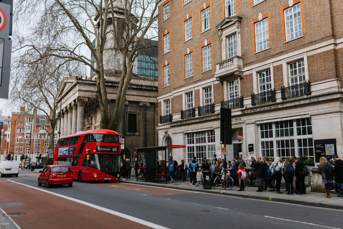 Crowd of people waiting at a bus stop near a historic building with a red double-decker bus and a red car on the street in London.