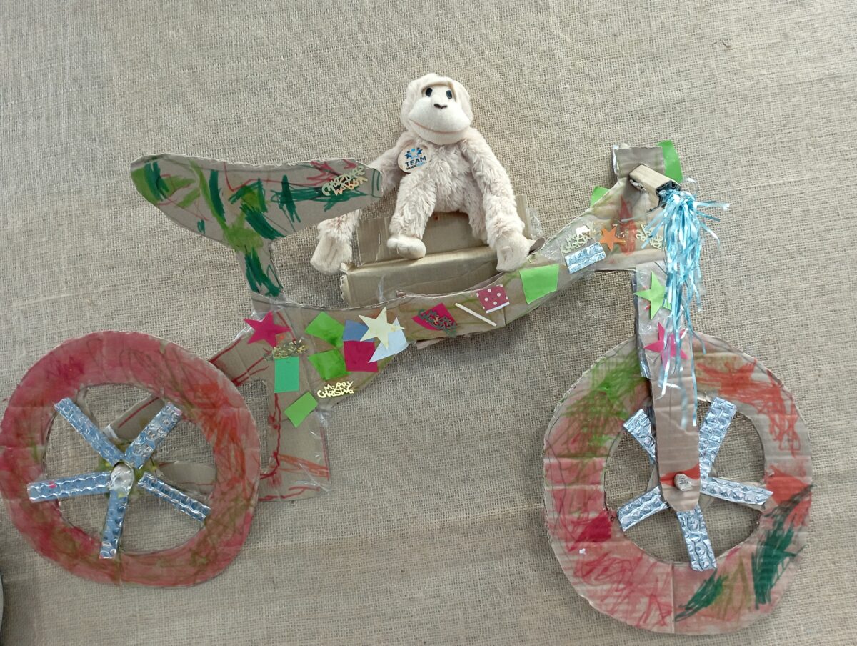 Handmade bicycle model crafted from cardboard and decorated with colorful paper and foil, featuring a plush white monkey seated on top.