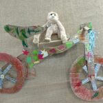 Handmade bicycle model crafted from cardboard and decorated with colorful paper and foil, featuring a plush white monkey seated on top.