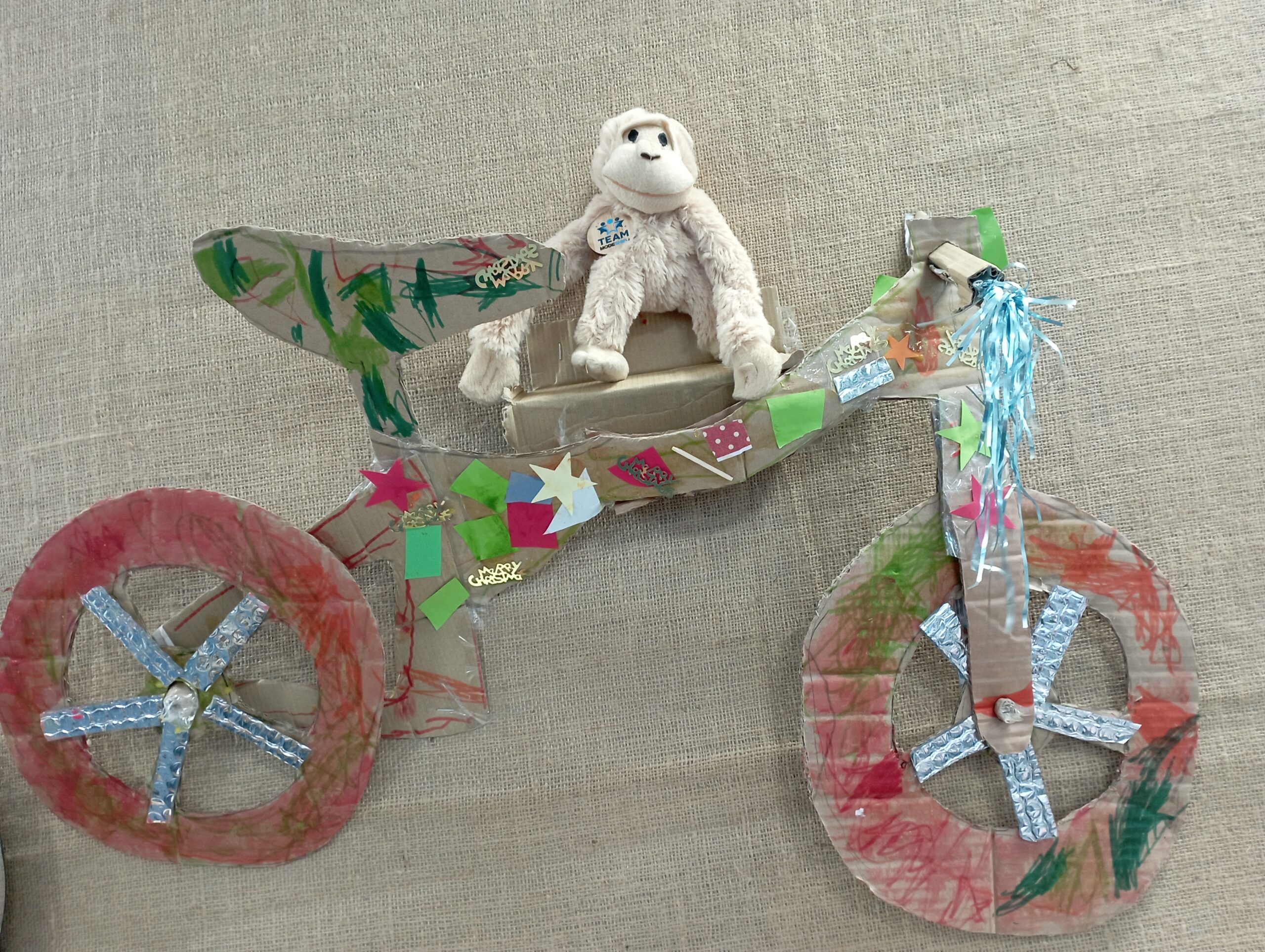 Handmade bicycle model crafted from cardboard and decorated with colorful paper and foil, featuring a plush white monkey seated on top.