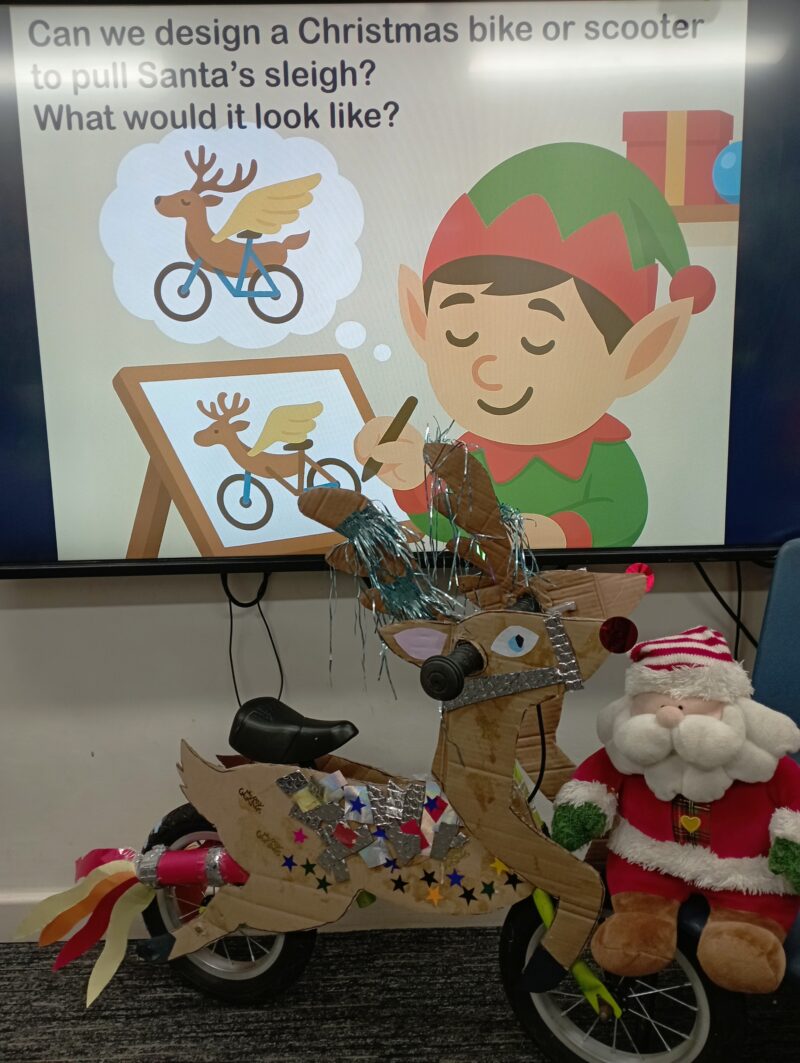 A cardboard reindeer bike decorated with stars and tinsel, designed to pull Santa's sleigh, displayed next to a Santa plush toy.