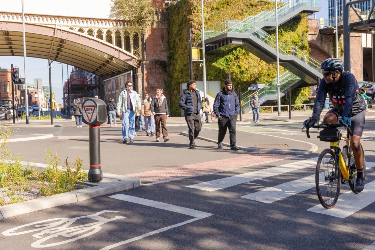 Cyclist wearing a helmet rides on a marked bike lane crossing a street with pedestrians walking nearby under a metal bridge in an urban area.