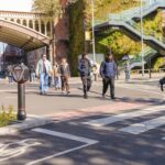 Cyclist wearing a helmet rides on a marked bike lane crossing a street with pedestrians walking nearby under a metal bridge in an urban area.