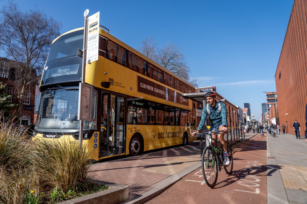 Yellow double-decker bus stopped at a city bus stop with a cyclist riding on a dedicated cycle lane beside it under a clear blue sky.