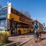 Yellow double-decker bus stopped at a city bus stop with a cyclist riding on a dedicated cycle lane beside it under a clear blue sky.