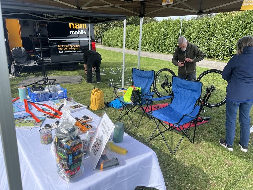 Outdoor setup with a table of cycling accessories and two blue folding chairs under a canopy near a mobile service van.