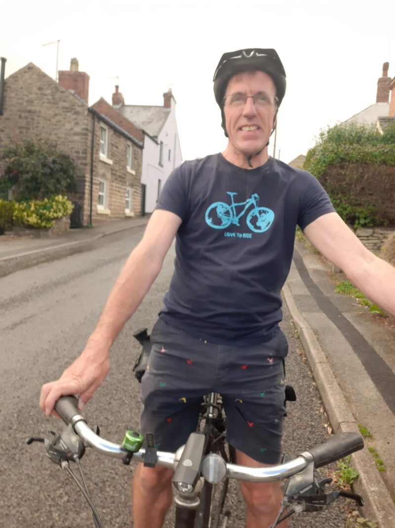 Person wearing a helmet and a navy blue t-shirt with a bicycle graphic, standing with hands on the handlebars of a bike on a quiet village street.