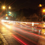 Long exposure of a busy street at night showing streaks of red and white car lights under green traffic signals.