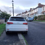 White Audi parked partially on a sidewalk in a residential neighborhood with terraced houses lining the street.