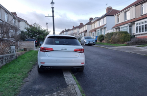 White Audi parked partially on a sidewalk in a residential neighborhood with terraced houses lining the street.