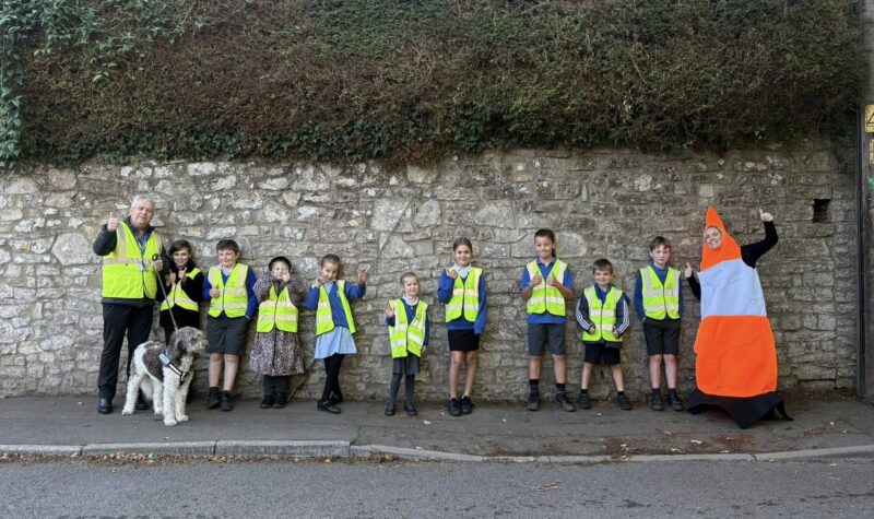 Group of children and an adult wearing yellow safety vests standing on a pavement by a stone wall, with one person dressed in an orange and white traffic cone costume.