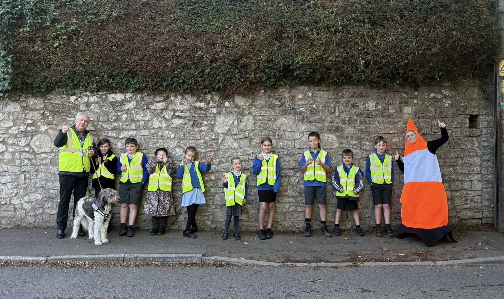 Group of children and an adult wearing yellow safety vests standing on a pavement by a stone wall, with one person dressed in an orange and white traffic cone costume.
