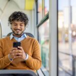 Person wearing a brown sweater sitting on a green bus seat, focused on their smartphone during a daytime ride.