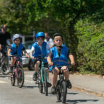 Three children wearing blue safety vests and helmets ride bicycles on a sunny street, followed by adults on bikes in a green, leafy neighborhood.