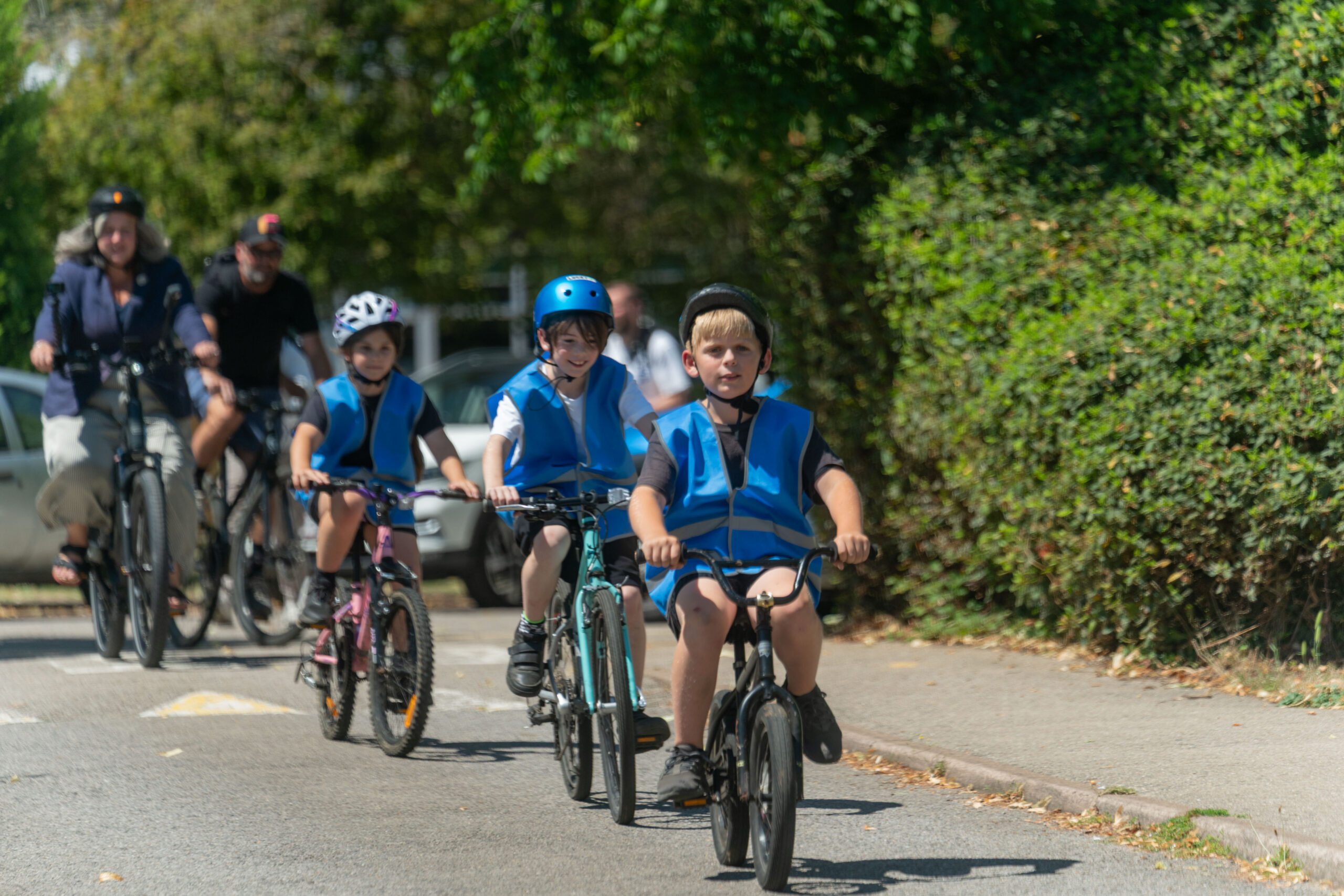 Three children wearing blue safety vests and helmets ride bicycles on a sunny street, followed by adults on bikes in a green, leafy neighborhood.