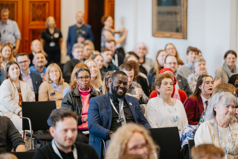 A diverse audience of people are seated in chairs facing forward, listening to a presentation or speaker in a conference or event hall.
