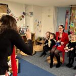 Woman taking a photo of a woman and three children holding stuffed animals in a colorful classroom.