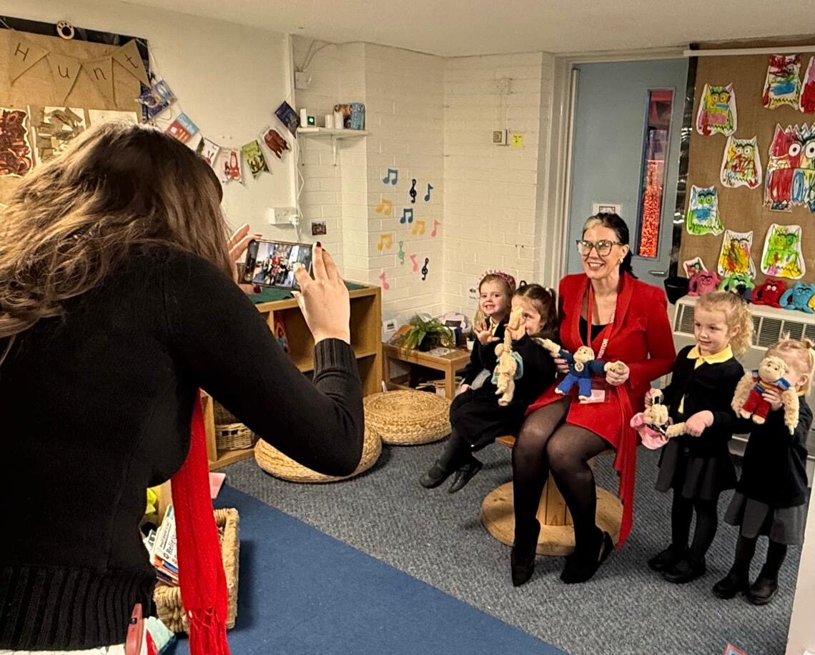 Woman taking a photo of a woman and three children holding stuffed animals in a colorful classroom.