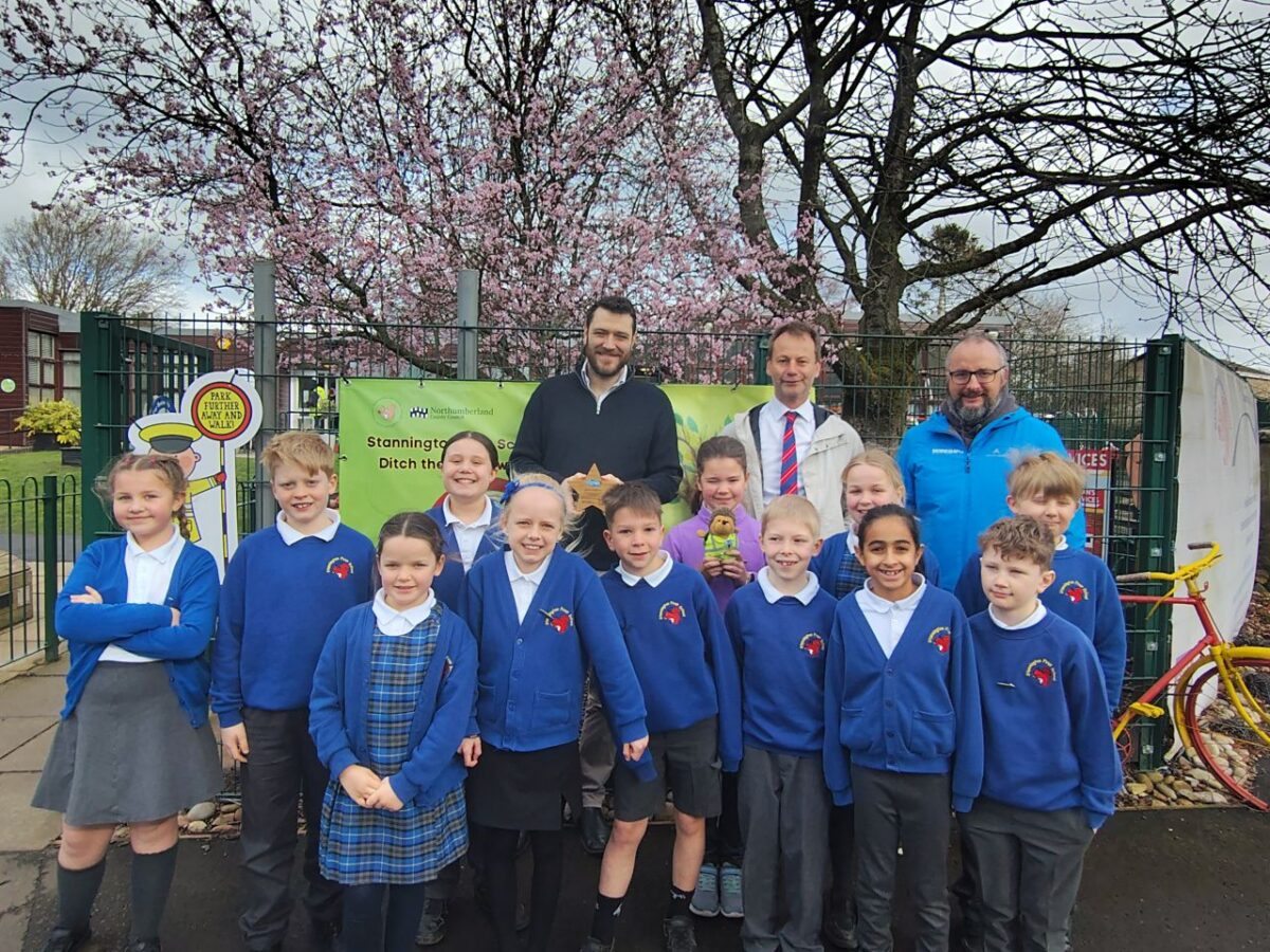 Group of schoolchildren in blue uniforms posing outdoors with two adults under blooming cherry trees near a school entrance.