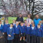 Group of schoolchildren in blue uniforms posing outdoors with two adults under blooming cherry trees near a school entrance.