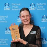 Woman in black dress holding a wooden National Travel Awards trophy against a blue branded backdrop.