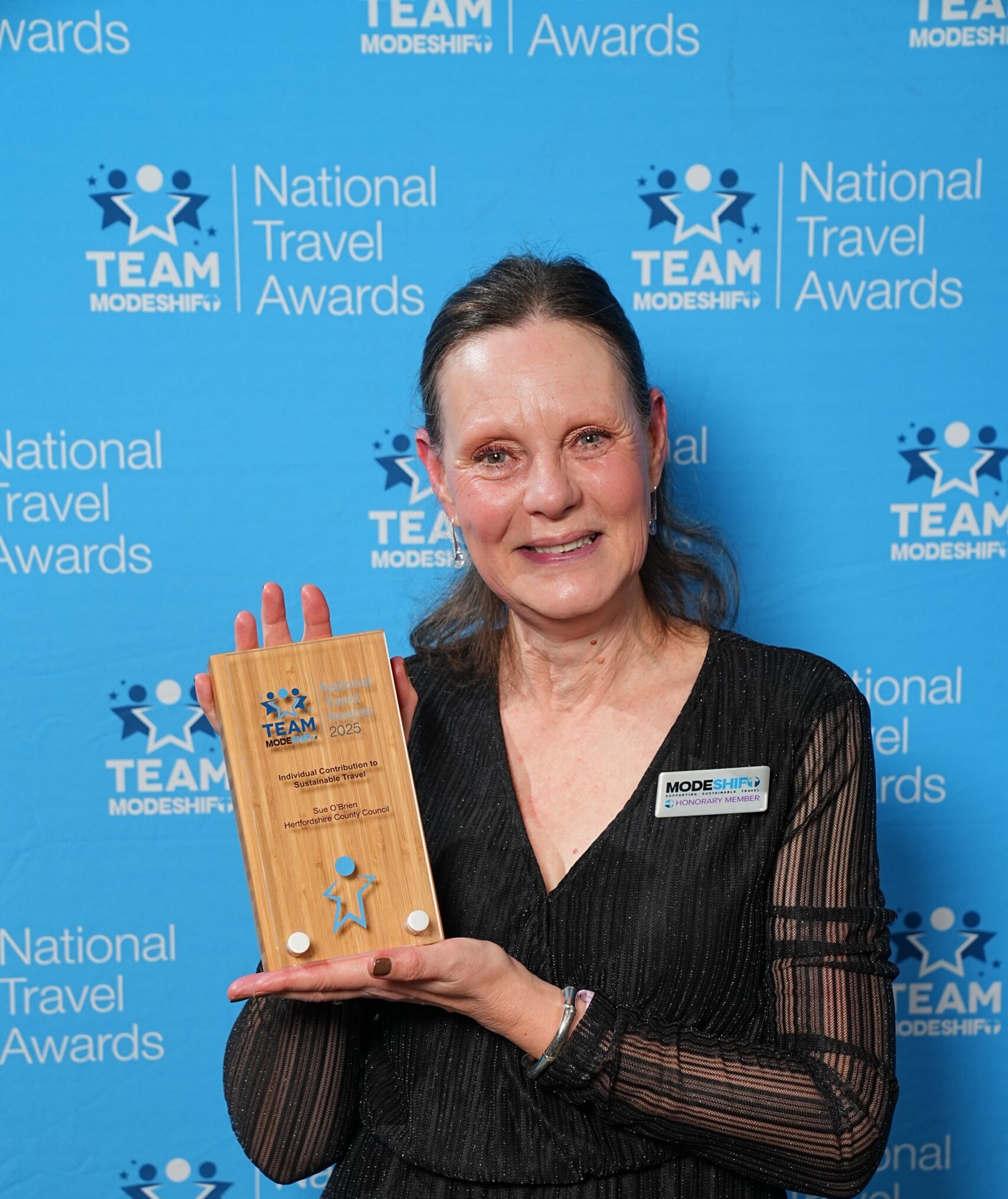 Woman in black dress holding a wooden National Travel Awards trophy against a blue branded backdrop.