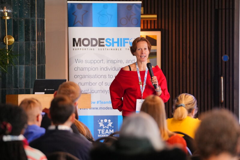 A woman in a red off-the-shoulder top speaks into a microphone at a podium, standing in front of a banner for "MODESHIFT SUPPORTING SUSTAINABLE TRAVEL" to an audience.