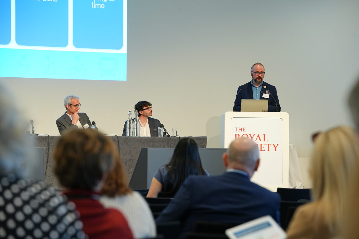 A speaker stands at a podium at a conference in The Royal Society, presenting to an audience. Behind him, a large screen displays “Modeshift National STARS Summit – Celebrating Sustainable Travel Excellence, London 18 March 2026.” Three panelists sit at a table beside him with microphones, and a Modeshift-branded banner with photos and logos is visible to the left.