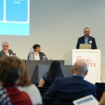 A speaker stands at a podium at a conference in The Royal Society, presenting to an audience. Behind him, a large screen displays “Modeshift National STARS Summit – Celebrating Sustainable Travel Excellence, London 18 March 2026.” Three panelists sit at a table beside him with microphones, and a Modeshift-branded banner with photos and logos is visible to the left.