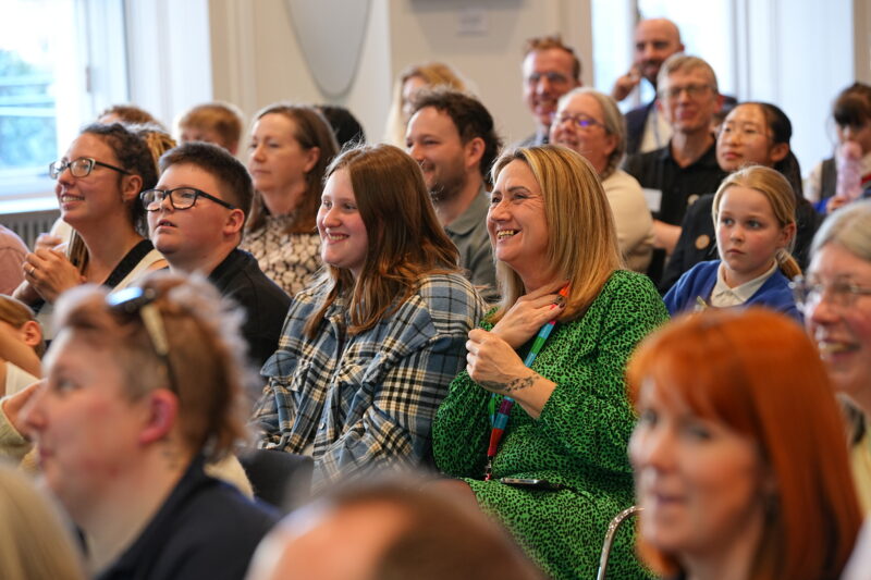 A diverse group of people, ranging in age from children to adults, are seated and looking forward, some with expressions of joy and interest, at an event or presentation. One woman in the center has a green patterned dress and is smiling broadly. Several others are visible in the foreground and background, with a variety of facial expressions and attire, including glasses and plaid shirts. The lighting suggests an indoor setting.