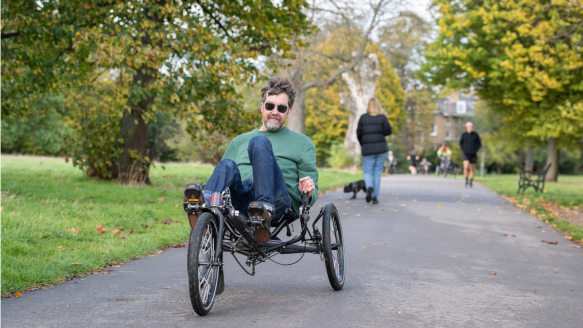 Person riding a three-wheeled recumbent bike on a paved park path surrounded by green and autumnal trees.