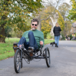Person riding a three-wheeled recumbent bike on a paved park path surrounded by green and autumnal trees.