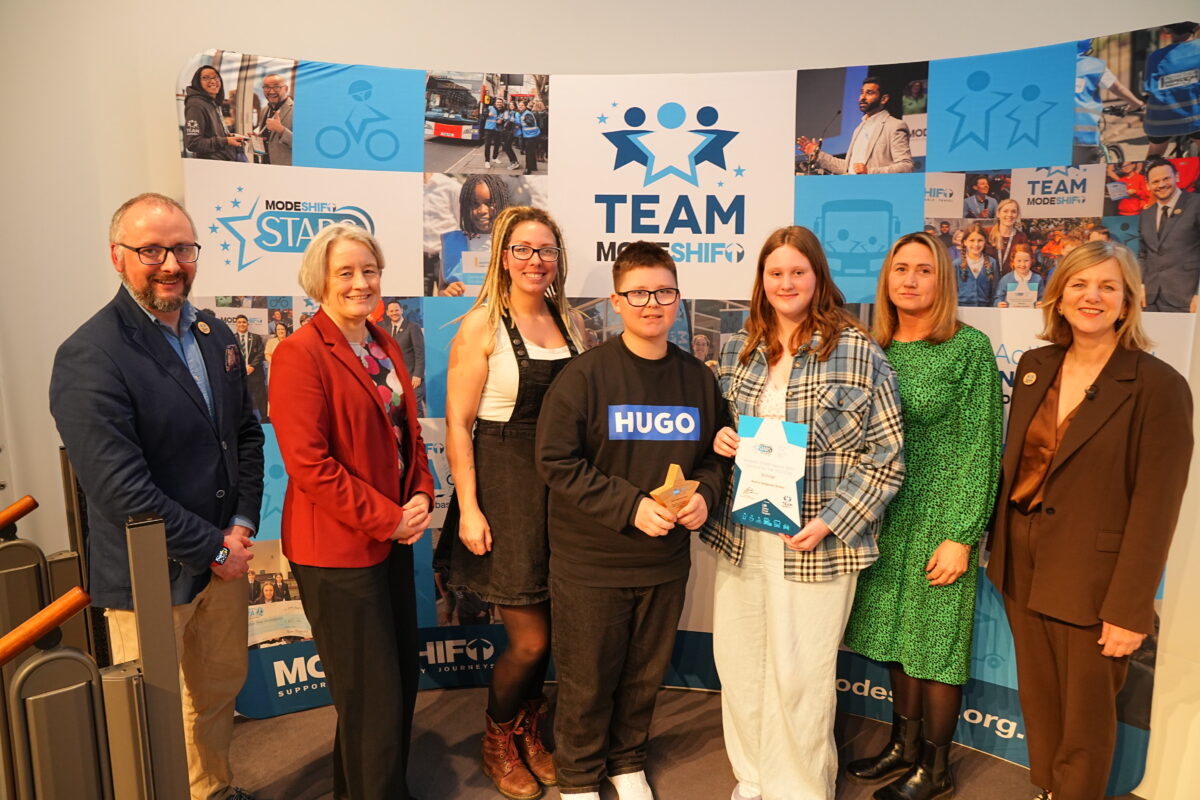 Group of seven people posing in front of a blue banner promoting "Team ModeShift" and "ModeShift Stars". A teenage boy and a teenage girl stand in the center holding an award and a certificate, both smiling, with a banner behind them showing "winner." The others are smiling and standing to the left and right sides of the two. The boy wears a black shirt with "Hugo" on the front. The girl wears a plaid shirt and white pants. Other people are wearing business casual clothes, some with jackets.