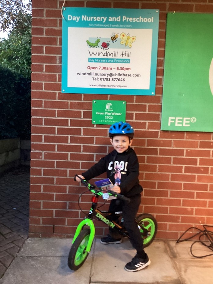 Child wearing a blue helmet rides a green balance bike outside Windmill Hill Day Nursery and Preschool building.