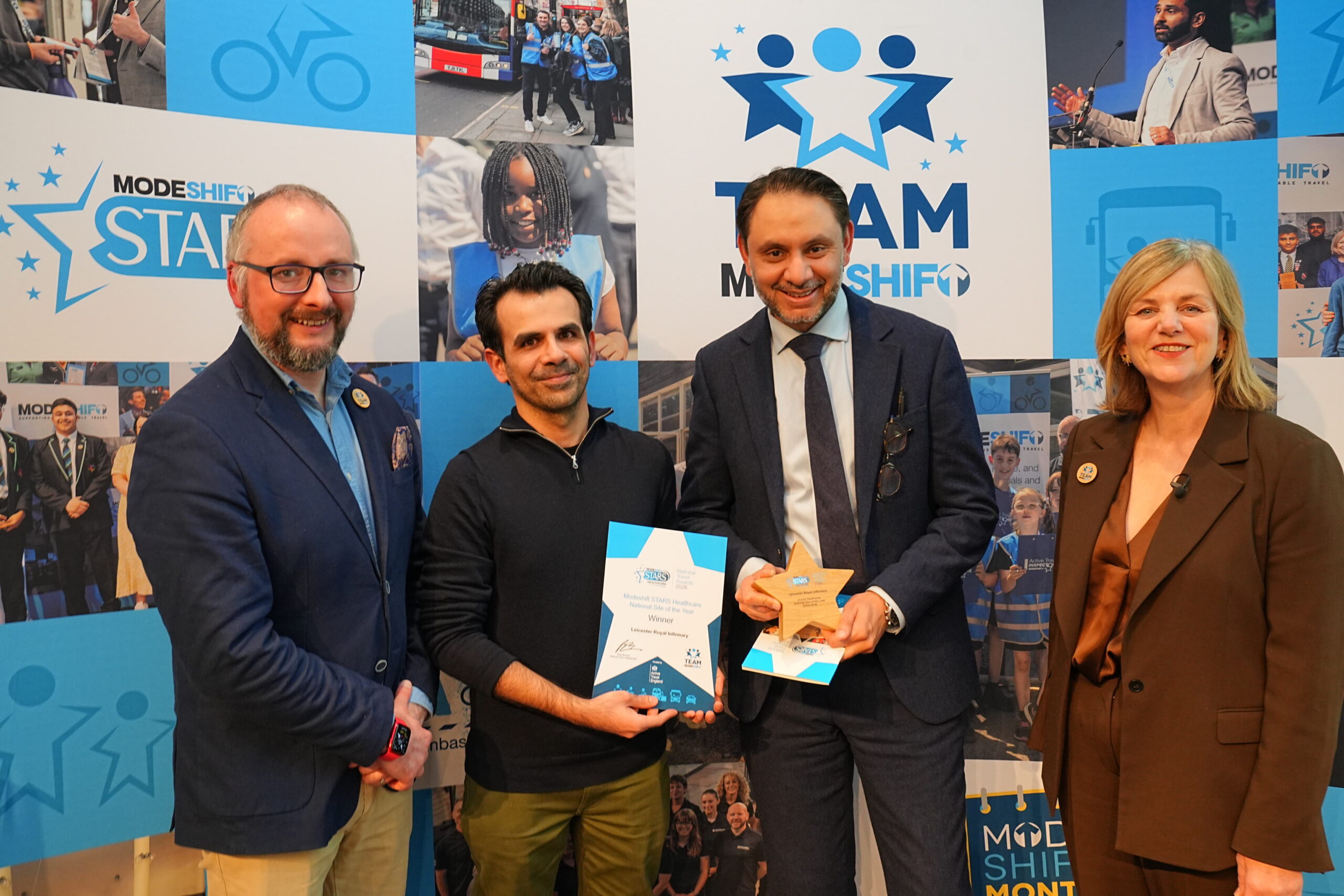 Four people standing in front of a blue and white backdrop with the text "TEAM MODESHIFT". Two men are holding awards. The man on the left is holding a certificate that reads "Modeshift STARS Healthcare, National Site of the Year, Winner, Leicester Royal Infirmary". The man in the middle holds a star-shaped wooden award. All four people are smiling and appear to be at an awards ceremony. The backdrop displays the logo "MODESHIFT STARS".