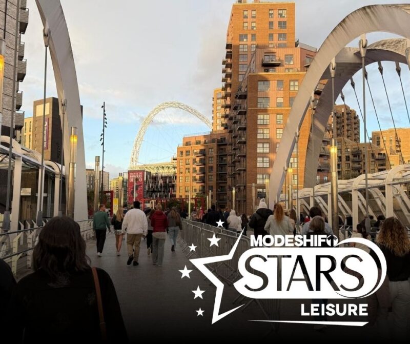 A crowd of people walk across a pedestrian bridge towards the iconic arch of Wembley Stadium in London, with modern apartment buildings on either side and a graphic overlay for "MODESHIFT STARS LEISURE."