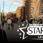 A crowd of people walk across a pedestrian bridge towards the iconic arch of Wembley Stadium in London, with modern apartment buildings on either side and a graphic overlay for "MODESHIFT STARS LEISURE."