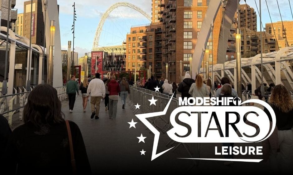 A crowd of people walk across a pedestrian bridge towards the iconic arch of Wembley Stadium in London, with modern apartment buildings on either side and a graphic overlay for "MODESHIFT STARS LEISURE."