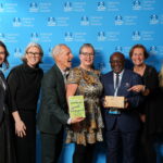 A group of seven people smiling and posing together at the National Travel Awards, standing in front of a blue branded backdrop. Two individuals at the centre hold an award plaque and a book titled “Potholes and Pavements”, while others stand close by, celebrating and pointing toward the award. Everyone is dressed in smart or business attire, creating a formal yet cheerful atmosphere.