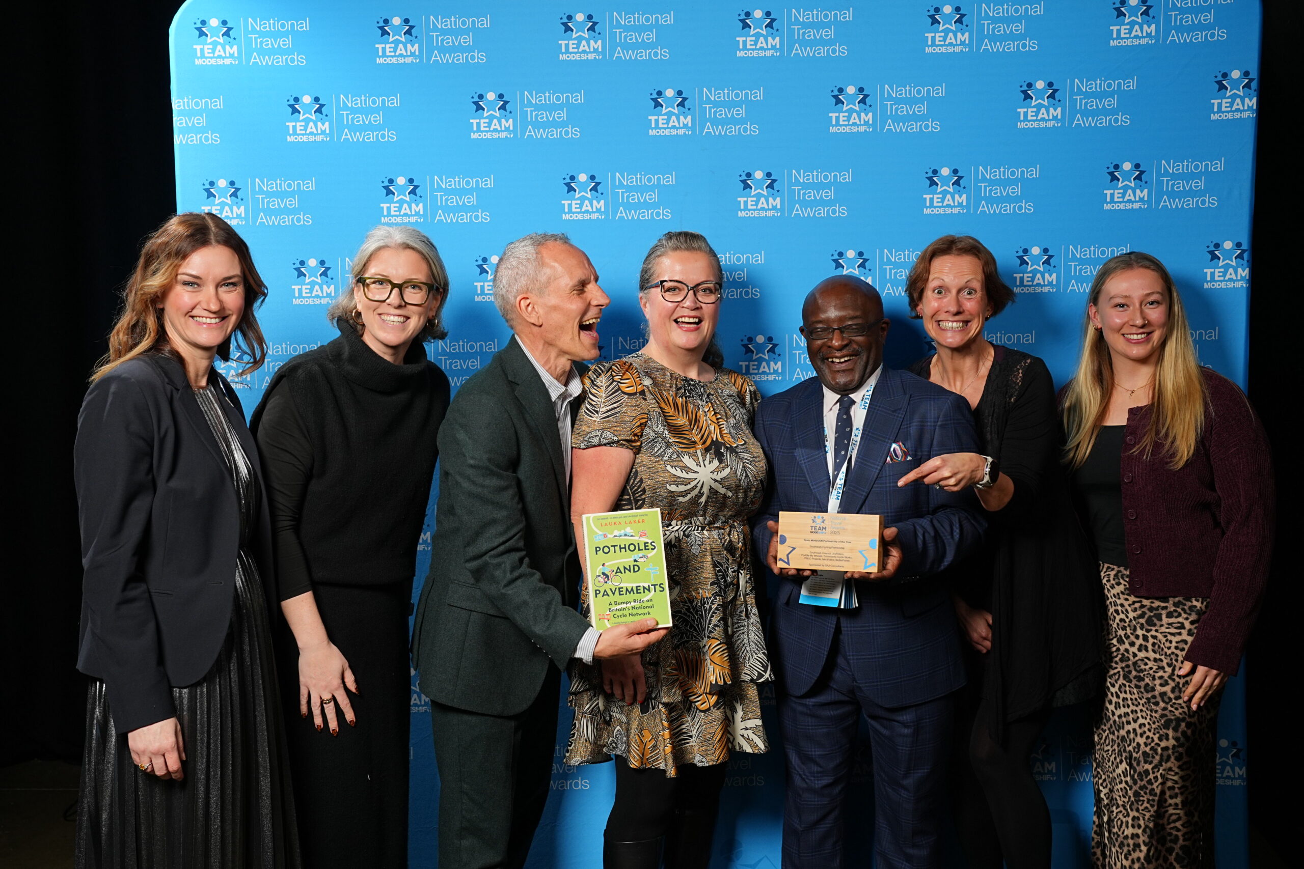 A group of seven people smiling and posing together at the National Travel Awards, standing in front of a blue branded backdrop. Two individuals at the centre hold an award plaque and a book titled “Potholes and Pavements”, while others stand close by, celebrating and pointing toward the award. Everyone is dressed in smart or business attire, creating a formal yet cheerful atmosphere.
