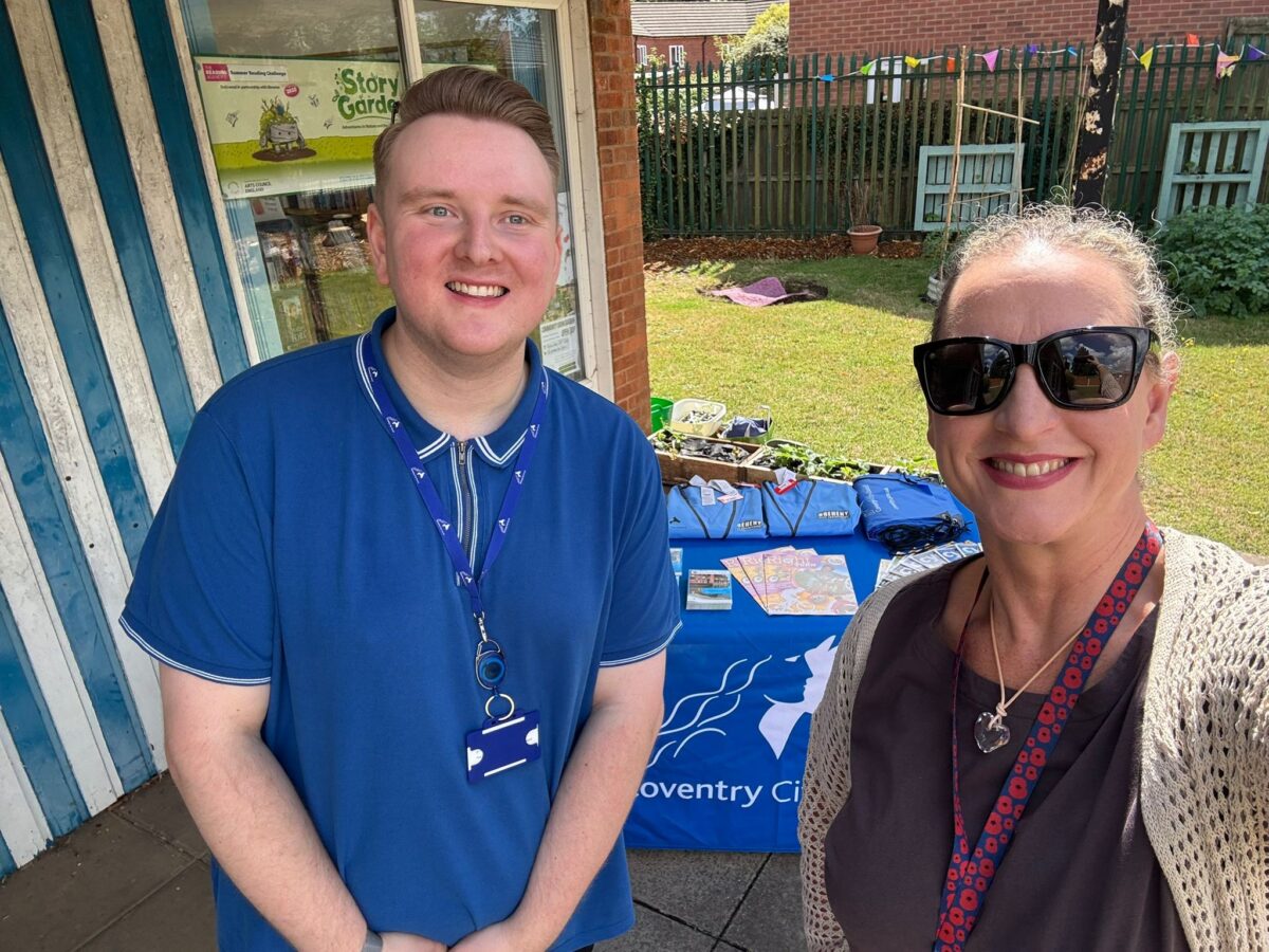 Two people standing outside near a table covered with blue cloth and promotional materials in a garden setting.