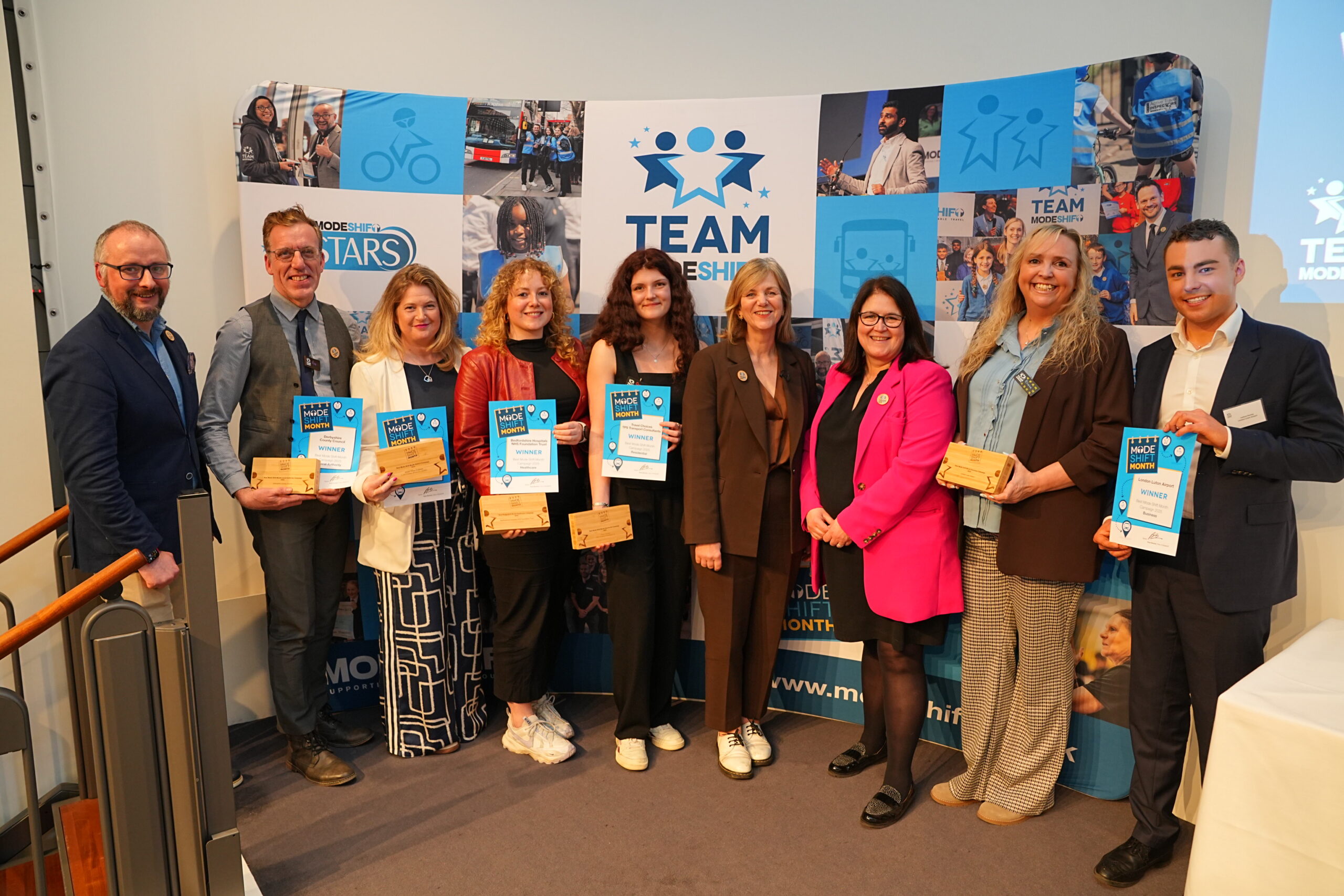 A group of nine people, men and women, smiling and holding certificates and wooden awards at an event with a backdrop featuring the "Mode Shift Month" logo and imagery. The individuals are dressed in business attire and are positioned in front of a backdrop displaying "Team Mode Shift" and other related graphics.