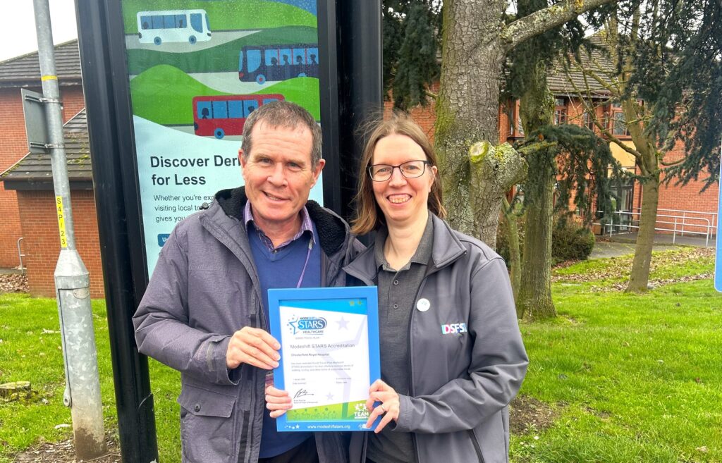Two people standing outdoors holding a framed certificate with a bus stop and green grass in the background. (L to R) Michael Reardon, Senior Transport Officer – Derbyshire County Council, Sally Ludditt, , Senior Health, Safety & Environmental Advisor, DSFS